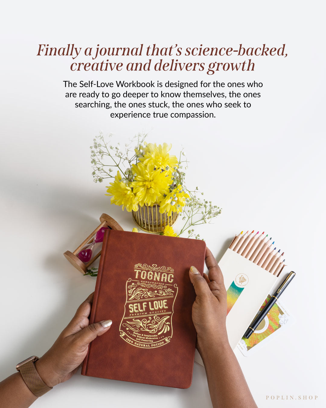Brown journal with 'Self Love' design held by hands, surrounded by yellow flowers and stationery on a white surface.