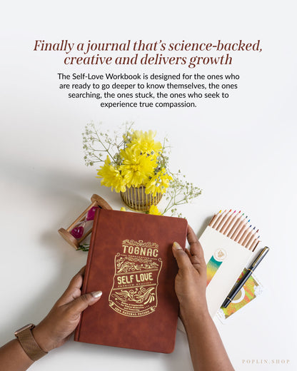 Brown journal with 'Self Love' design held by hands, surrounded by yellow flowers and stationery on a white surface.